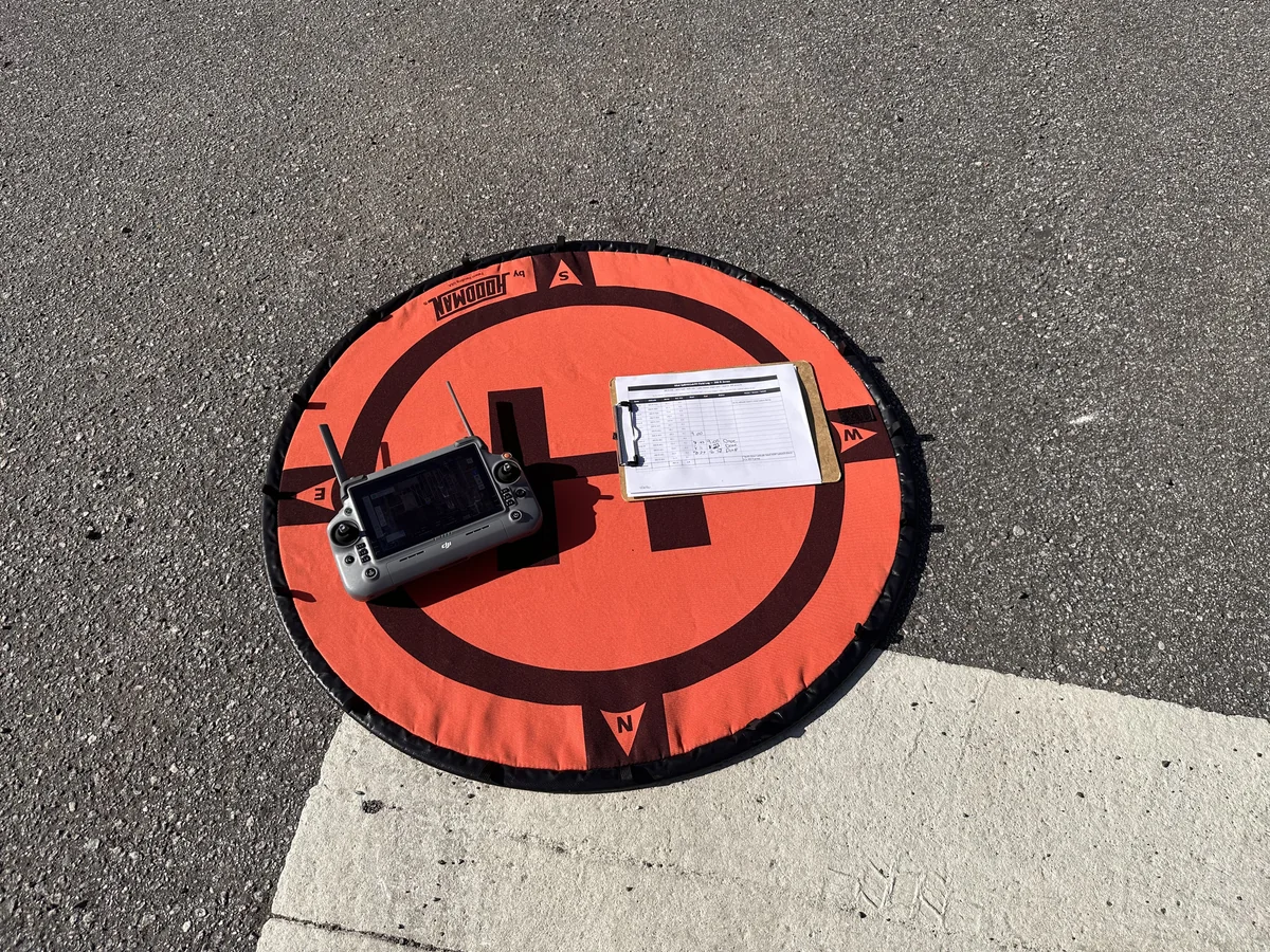 Top-down view of a Hoodman landing pad with the aircraft controller and a clipboard holding the printed field log at the launch site for a multi-zone aerial mapping mission