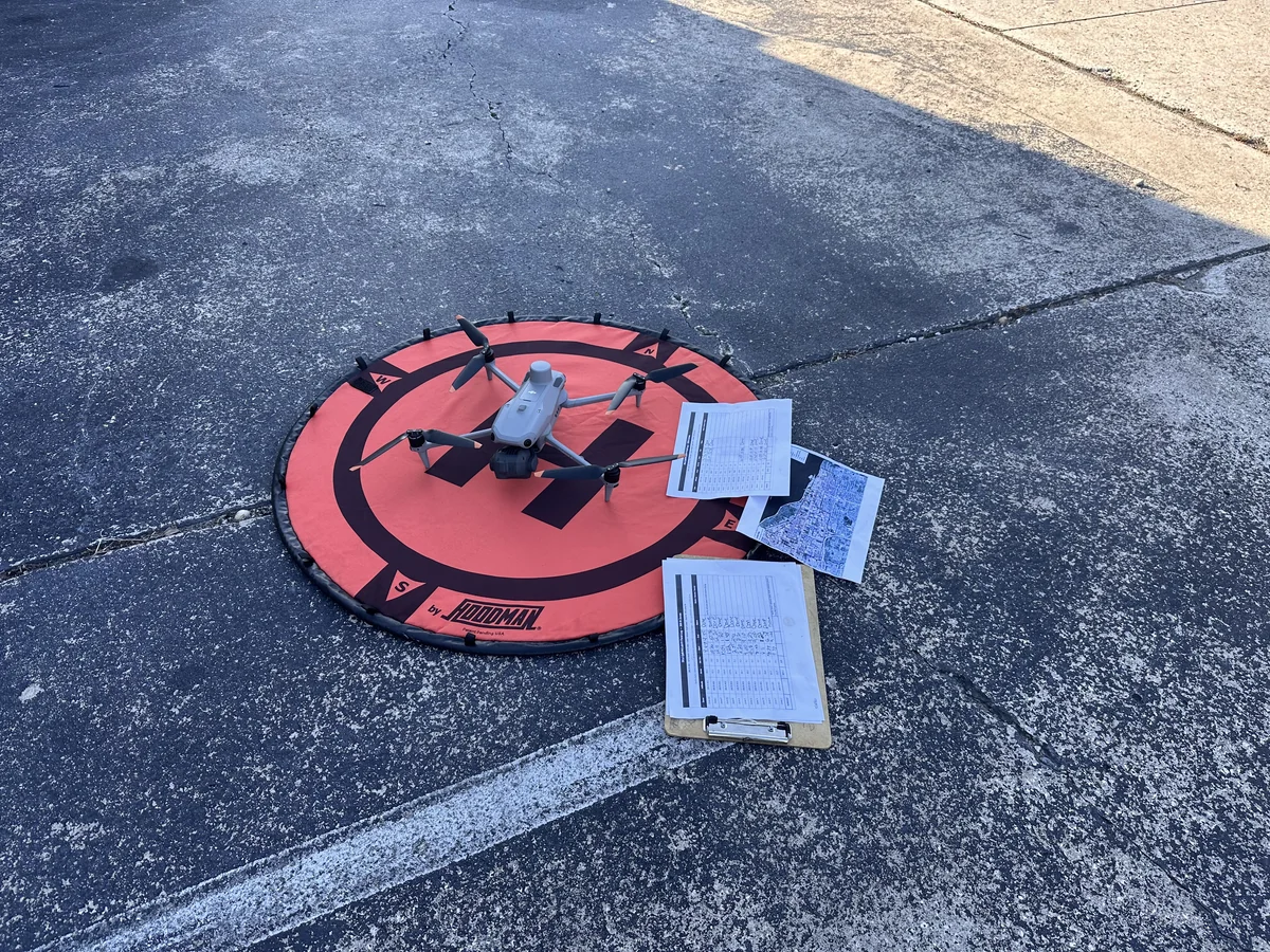 The field aircraft staged on a Hoodman landing pad with the printed field log, flight documentation, and a working aerial map staged on the adjacent pavement at the launch site for a multi-zone aerial mapping mission