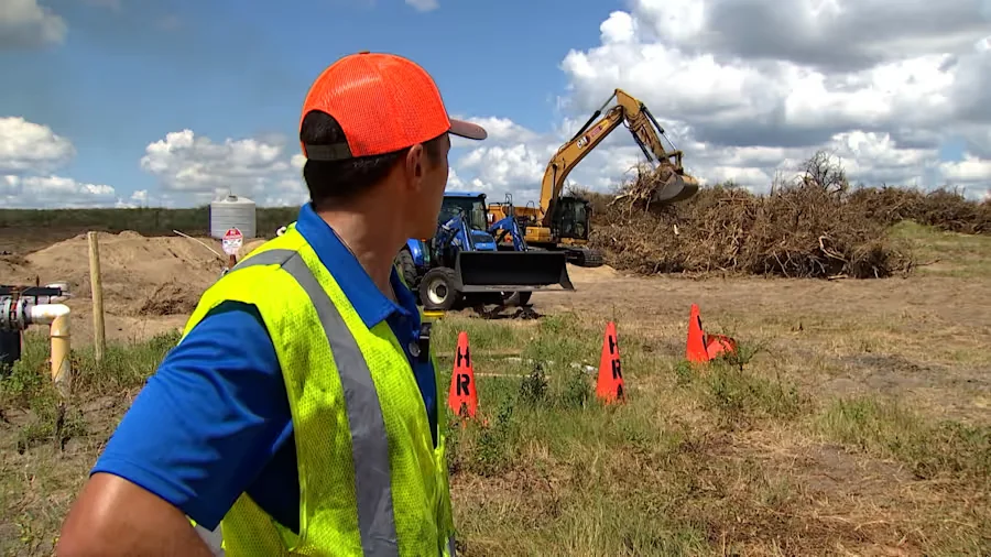 Site supervisor in an orange cap and high-visibility yellow safety vest observes a yellow Caterpillar excavator and blue tractor sorting a large pile of woody citrus brush at a Central Florida burn site, with safety cones and a portable water tank staged nearby