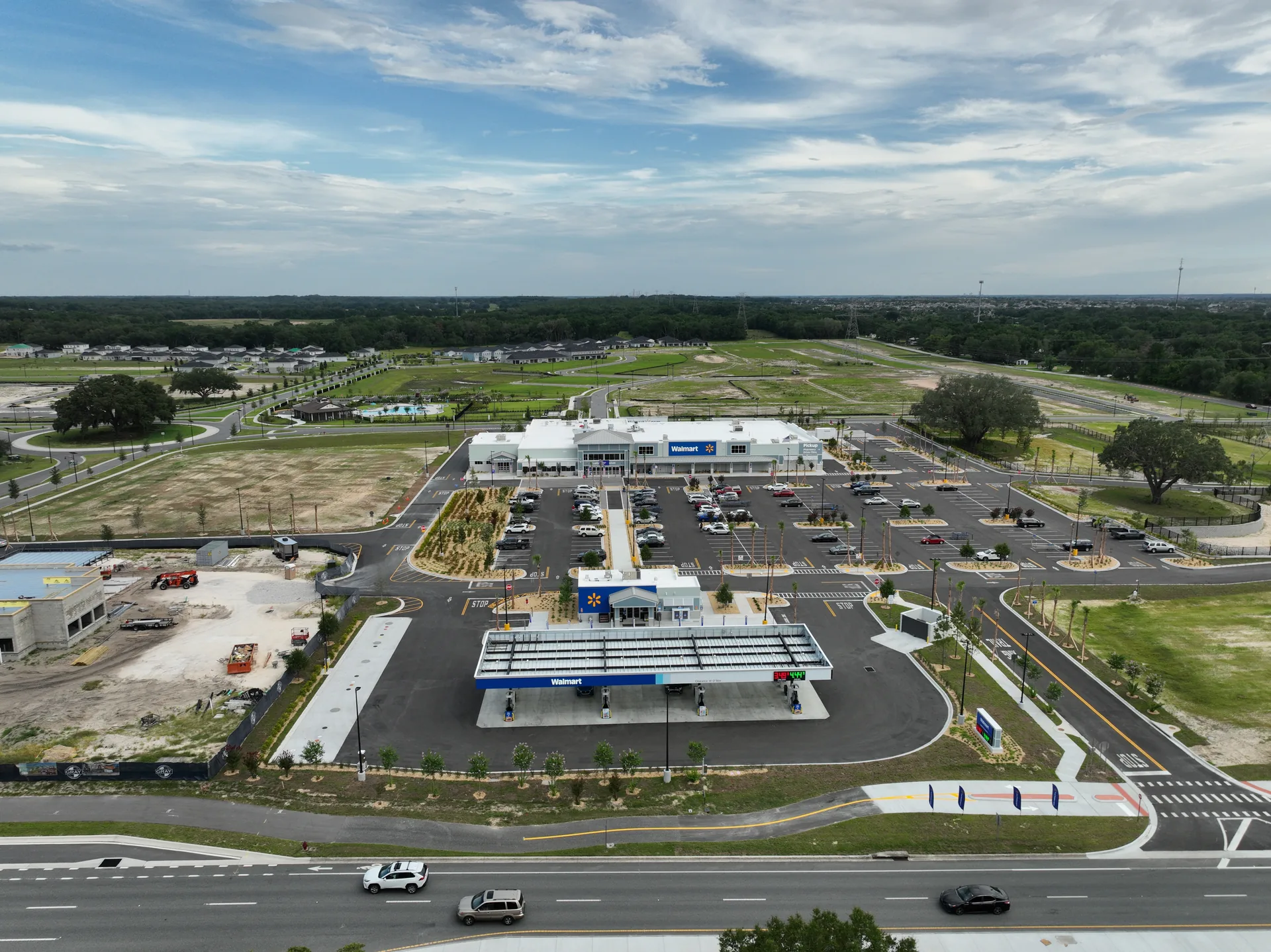 Aerial drone view of the completed Walmart Supercenter with gas station fueling canopy at 1204 Crabapple Lane, Lady Lake, Florida, captured at the end of an 11-month weekly aerial documentation engagement in June 2026. The image shows the operational Walmart storefront, the fueling canopy with gas pumps, the freshly striped parking lot with newly planted palm trees, an active outparcel CMU building under construction on the left edge, and the surrounding residential community and access roads in their final configuration.