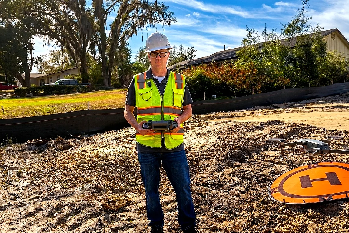 Chad Hammond, founder of Skyland Pixels, on a Central Florida construction site in white hard hat and high-visibility safety vest, operating a DJI drone with remote controller, construction site and DJI Mavic drone visible